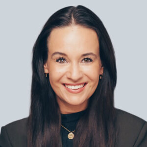 Eine Frau mit langen, glatten dunklen Haaren lächelt in die Kamera. Sie trägt ein schwarzes Oberteil und einen Blazer, kleine Ohrringe und eine goldene Halskette mit Anhänger, die Professionalität in Strategie- und Transformationsberatung vor einem hellgrauen Hintergrund widerspiegeln.