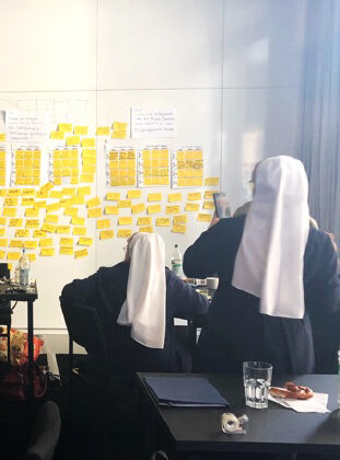 Zwei Nonnen in weißen Kitteln arbeiten an einem Tisch vor einer Wand, die mit vielen gelben Zetteln und Papieren bedeckt ist. Der Raum ist hell erleuchtet, auf dem Tisch stehen Notizbücher, Wasserflaschen und Snacks.