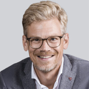 Ein lächelnder Mann mit blondem Haar, einem kurzen Bart und einer Brille trägt einen hellblauen Blazer über einem weißen Hemd und posiert vor einem schlichten hellen Hintergrund.