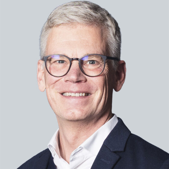 Ein lächelnder Mann mit blondem Haar, einem kurzen Bart und einer Brille trägt einen hellblauen Blazer über einem weißen Hemd und posiert vor einem schlichten hellen Hintergrund.