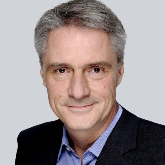 Ein Mann mittleren Alters mit kurzen grauen Haaren lächelt freundlich. Er trägt eine dunkle Anzugsjacke über einem hellblauen Hemd mit Kragen. Der Hintergrund ist schlicht und hellgrau.