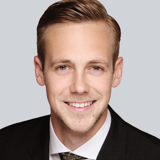 Ein junger Mann mit kurzen hellbraunen Haaren, der einen dunklen Anzug, ein weißes Hemd und eine gemusterte Krawatte trägt, lächelt in die Kamera vor einem schlichten hellgrauen Hintergrund.