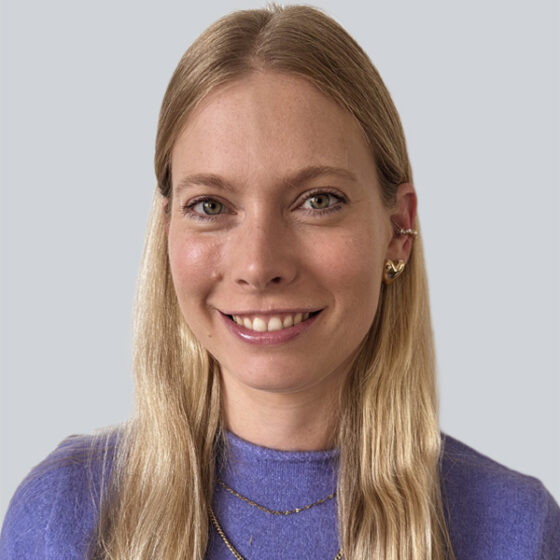 Eine Frau mit langen blonden Haaren, die ein lila Oberteil, goldene Ohrringe und eine mehrlagige goldene Halskette trägt, lächelt in die Kamera vor einem schlichten hellgrauen Hintergrund.