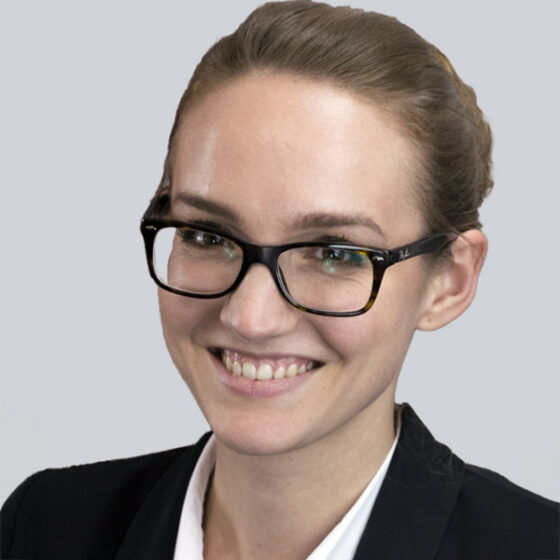 Eine Frau mit heller Haut, braunem, nach hinten gestecktem Haar, schwarzer Brille, weißem Hemd und dunklem Blazer lächelt in die Kamera vor einem schlichten hellgrauen Hintergrund.