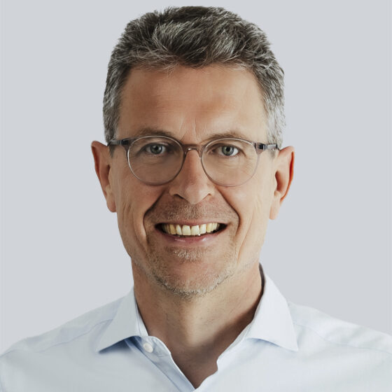 Ein lächelnder Mann mittleren Alters mit kurzen grauen Haaren, der eine Brille und ein hellblaues Hemd mit Kragen trägt, posiert vor einem schlichten hellgrauen Hintergrund.