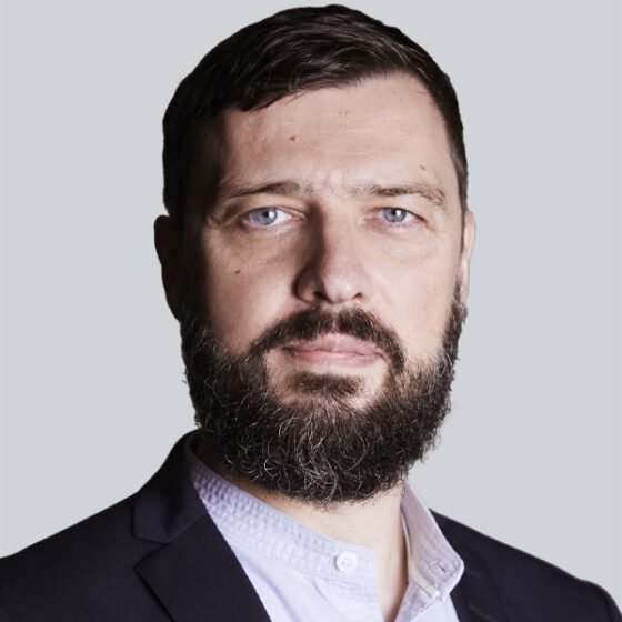 Ein Mann mit kurzen braunen Haaren, Vollbart und blauen Augen, der einen dunklen Blazer über einem weißen Button-up-Hemd trägt, posiert vor einem schlichten hellgrauen Hintergrund.