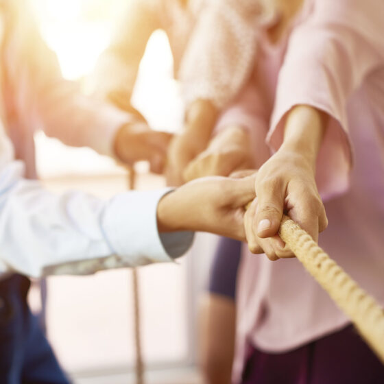 Nahaufnahme von mehreren Personen, die beim Tauziehen an einem Seil ziehen, wobei sie das Seil mit den Händen fest umklammern. Der Hintergrund ist hell und unscharf.