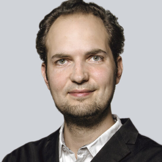 Ein Mann mit kurzen, lockigen braunen Haaren und heller Haut, der einen schwarzen Blazer über einem weißen Hemd mit Kragen trägt, lächelt in die Kamera vor einem schlichten hellgrauen Hintergrund.