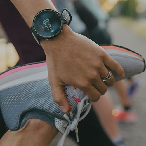 Eine Person, die eine Smartwatch trägt, streckt ihr Bein, indem sie den Fuß mit dem Schuh hinter sich hält, um sich auf einen Lauf oder ein Training vorzubereiten. Die Uhr zeigt die Zeit von 8:35 an.