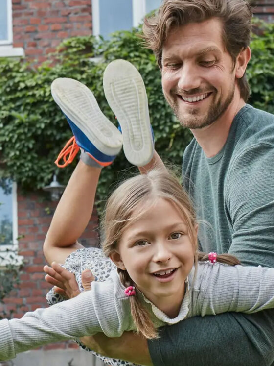 Ein lächelnder Mann hebt ein lachendes junges Mädchen mit Zöpfen hoch, das seine Arme wie ein Flugzeug ausstreckt. Sie stehen im Freien vor einem Haus mit Backsteinmauern und grünem Efeu.