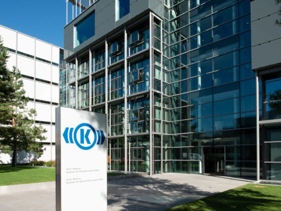 Modernes gläsernes Bürogebäude mit einem Schild an der Vorderseite, das ein blaues kreisförmiges Logo und Text zeigt. Bäume und eine Rasenfläche sind zu sehen, und der Eingang ist mit großen Fenstern und Stahlrahmen versehen.