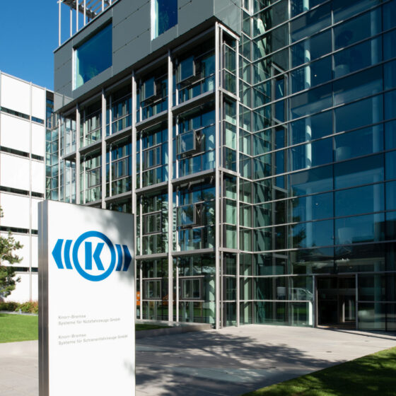 Modernes gläsernes Bürogebäude mit einem Schild an der Vorderseite, das ein blaues kreisförmiges Logo und Text zeigt. Bäume und eine Rasenfläche sind zu sehen, und der Eingang ist mit großen Fenstern und Stahlrahmen versehen.