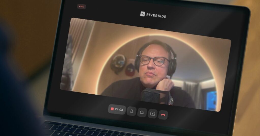 Ein Mann mit Brille und Kopfhörern spricht während eines Online-Gesprächs oder einer Aufzeichnung über die Erarbeitung von Zukunftsstrategie in ein Mikrofon, dargestellt auf einem Laptop-Bildschirm, auf dem die Oberfläche der Riverside-App sichtbar ist.