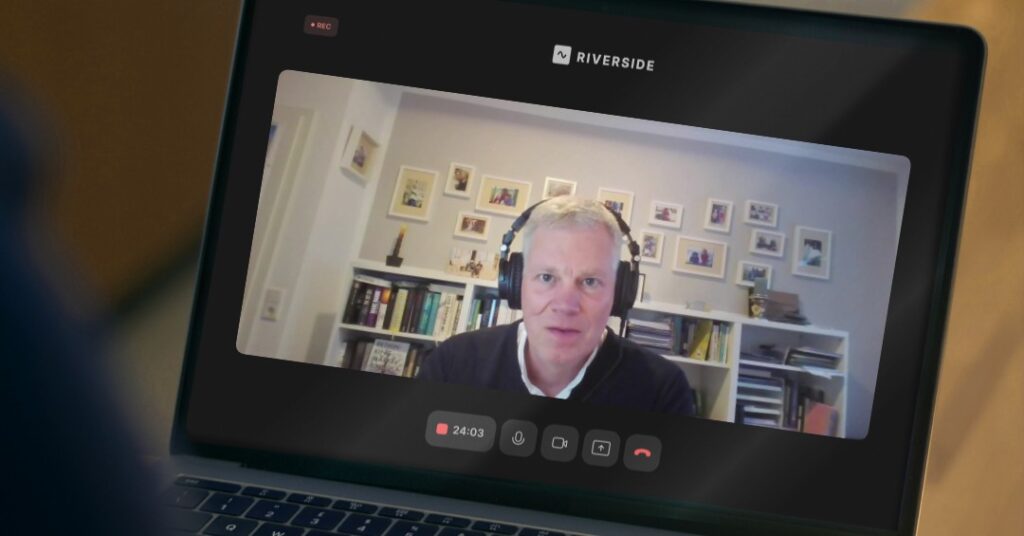 Ein Mann mit Kopfhörern sitzt in einem gut beleuchteten Raum mit Bücherregalen und gerahmten Fotos hinter sich und nimmt an einer virtuellen Riverside-Sitzung über die Erarbeitung von Zukunftsstrategie teil, die auf einem Laptop-Bildschirm angezeigt wird.