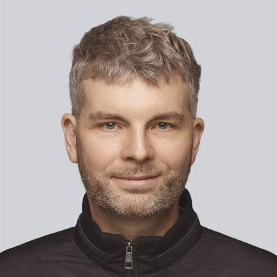 Porträt eines Mannes mit kurzem, hellbraunem Haar und gestutztem Bart, der eine schwarze Jacke mit Reißverschluss trägt. Er lächelt sanft und spiegelt seine Expertise in Strategie- und Transformationsberatung vor einem schlichten hellgrauen Hintergrund wider.