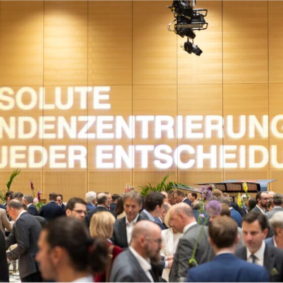 Eine große Gruppe von Menschen in Geschäftskleidung mischt sich bei einer Indoor-Veranstaltung von Knorr-Bremse. An der Wand steht in heller Schrift auf Deutsch: ABSOLUTE KUNDENZENTRIERUNG IN JEDER ENTSCHEIDUNG.