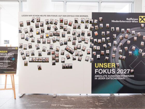 Auf einer Veranstaltung der Raiffeisen Niederösterreich-Wien steht neben einem großen Schild mit der Aufschrift UNSER FOKUS 2027 eine Schautafel mit vielen Sofortbildern, die an ähnliche Aufbauten auf Ausstellungen von Knorr-Bremse erinnert. Der Hintergrund ist hell und modern.