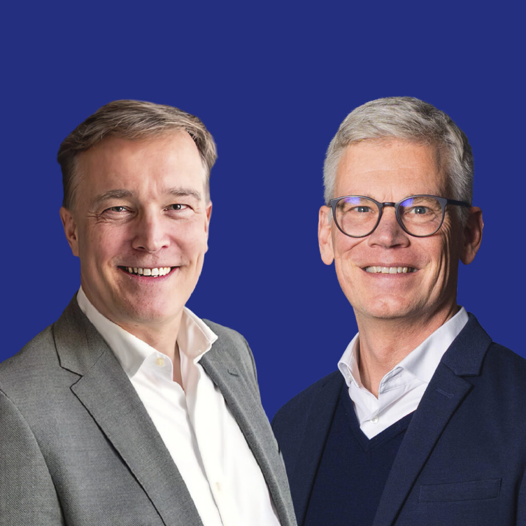 Zwei Männer in Business-Casual-Kleidung vor einem einfarbigen blauen Hintergrund, die über Rails Human Attention sprechen.